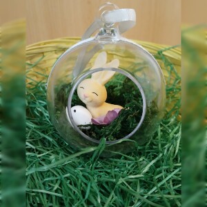 Handgefertigte, transparente Osterkugel aus Acryl mit einer Miniaturszene im Inneren: ein kleiner Osterhase sitzt auf einem Bett aus grünem Moos, daneben ein zartes Deko-Ei und feine Blüten. Die Kugel hängt an einem weißen Satinband und wirkt natürlich, frühlingshaft und elegant – ein stilvoller Osterschmuck zum Aufhängen oder Verschenken.