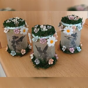 Handgefertigtes Feenglas mit romantischem Feenmotiv, verziert mit blauem Satinband, feinem Glitzerstaub und einer kleinen Mooslandschaft auf dem Deckel. Umgeben von zarten Kunstblumen wirkt das Glas wie ein leuchtender Miniaturgarten. Im Inneren sorgt ein flackerndes LED-Teelicht für warmes, magisches Licht – ein märchenhaftes Deko- und Nachtlicht für jeden Raum.