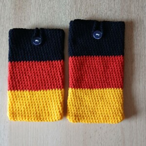 Handgehäkelte Handyhülle in den Farben Schwarz, Rot und Gold, angeordnet wie die deutsche Flagge. Aus robustem Baumwollgarn gefertigt, mit weicher Struktur und sicherem Knopfverschluss oben. Schützt das Smartphone vor Kratzern und Stößen. Erhältlich in zwei Größen – ein handgemachtes Accessoire für Fans und Liebhaber deutscher Farben.