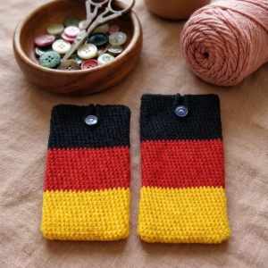 Handgehäkelte Handyhülle Deutschland Handyhülle in den Farben Schwarz, Rot und Gold, angeordnet wie die deutsche Flagge. Aus robustem Wollgarn gefertigt, mit weicher Struktur und sicherem Knopfverschluss oben. Schützt das Smartphone vor Kratzern und Stößen. Erhältlich in zwei Größen – ein handgemachtes Accessoire für Fans und Liebhaber deutscher Farben.