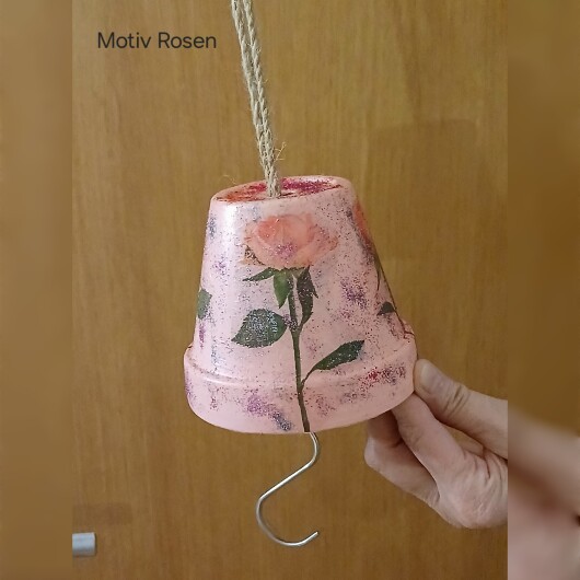 Handbemalte Futterglocke Motiv Rose aus einem Tontopf, bunt verziert mit Serviettentechnik, hängt an einer dicken Kordel. Im Inneren ist ein Metallhaken für Vogelfutter angebracht. Dekorative Winterfütterung für kleine Gartenvögel.