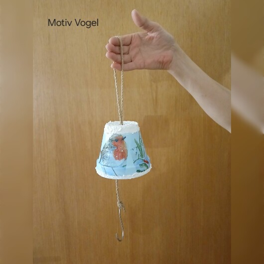Handbemalte Futterglocke Motiv Vogel aus einem Tontopf, bunt verziert mit Serviettentechnik, hängt an einer dicken Kordel. Im Inneren ist ein Metallhaken für Vogelfutter angebracht. Dekorative Winterfütterung für kleine Gartenvögel.