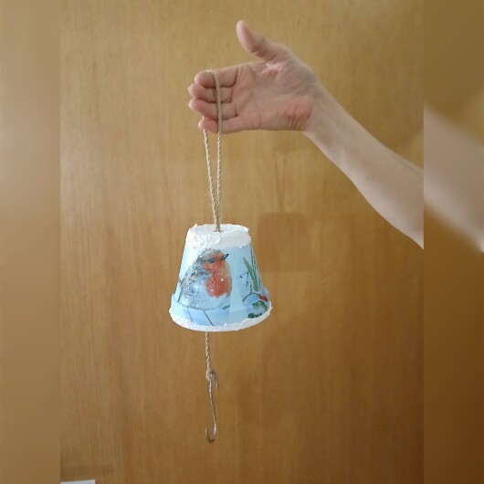 Handbemalte Futterglocke Motiv Vogel aus einem Tontopf, bunt verziert mit Serviettentechnik, hängt an einer dicken Kordel. Im Inneren ist ein Metallhaken für Vogelfutter angebracht. Dekorative Winterfütterung für kleine Gartenvögel.