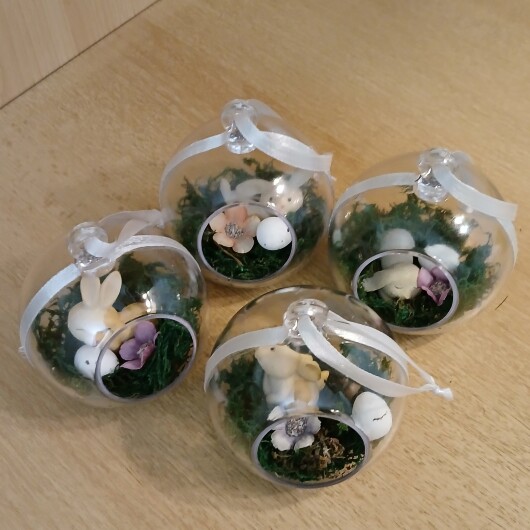 Handgefertigte, transparente Osterkugel aus Acryl mit einer Miniaturszene im Inneren: ein kleiner Osterhase sitzt auf einem Bett aus grünem Moos, daneben ein zartes Deko-Ei und feine Blüten. Die Kugel hängt an einem weißen Satinband und wirkt natürlich, frühlingshaft und elegant – ein stilvoller Osterschmuck zum Aufhängen oder Verschenken.