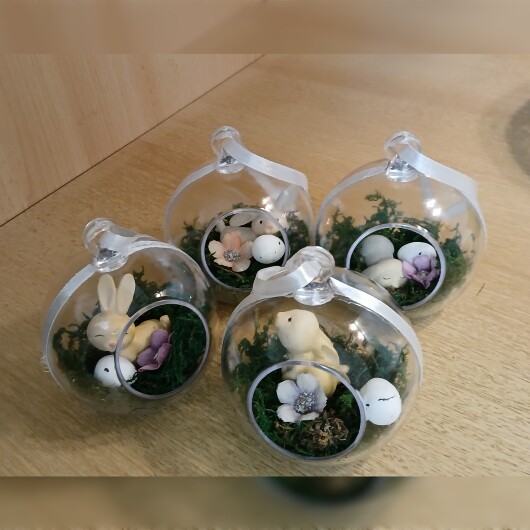 Handgefertigte, transparente Osterkugel aus Acryl mit einer Miniaturszene im Inneren: ein kleiner Osterhase sitzt auf einem Bett aus grünem Moos, daneben ein zartes Deko-Ei und feine Blüten. Die Kugel hängt an einem weißen Satinband und wirkt natürlich, frühlingshaft und elegant – ein stilvoller Osterschmuck zum Aufhängen oder Verschenken.