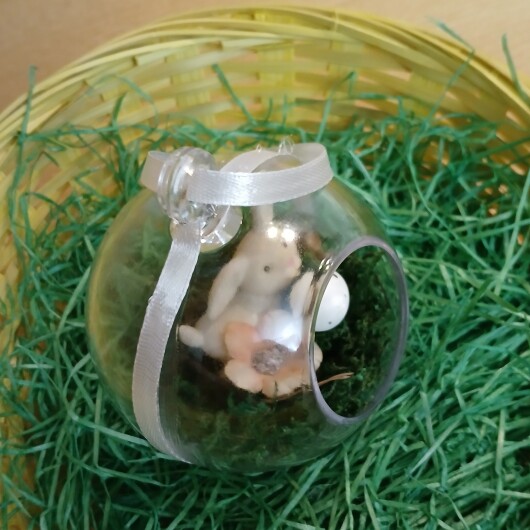 Handgefertigte, transparente Osterkugel aus Acryl mit einer Miniaturszene im Inneren: ein kleiner Osterhase sitzt auf einem Bett aus grünem Moos, daneben ein zartes Deko-Ei und feine Blüten. Die Kugel hängt an einem weißen Satinband und wirkt natürlich, frühlingshaft und elegant – ein stilvoller Osterschmuck zum Aufhängen oder Verschenken.