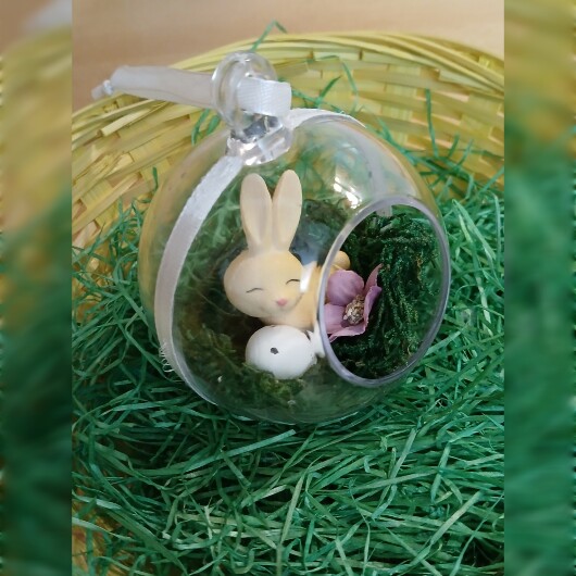 Handgefertigte, transparente Osterkugel aus Acryl mit einer Miniaturszene im Inneren: ein kleiner Osterhase sitzt auf einem Bett aus grünem Moos, daneben ein zartes Deko-Ei und feine Blüten. Die Kugel hängt an einem weißen Satinband und wirkt natürlich, frühlingshaft und elegant – ein stilvoller Osterschmuck zum Aufhängen oder Verschenken.