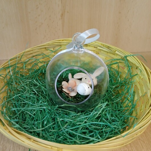 Handgefertigte, transparente Osterkugel aus Acryl mit einer Miniaturszene im Inneren: ein kleiner Osterhase sitzt auf einem Bett aus grünem Moos, daneben ein zartes Deko-Ei und feine Blüten. Die Kugel hängt an einem weißen Satinband und wirkt natürlich, frühlingshaft und elegant – ein stilvoller Osterschmuck zum Aufhängen oder Verschenken.