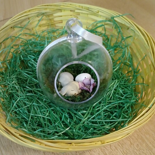 Handgefertigte, transparente Osterkugel aus Acryl mit einer Miniaturszene im Inneren: ein kleiner Osterhase sitzt auf einem Bett aus grünem Moos, daneben ein zartes Deko-Ei und feine Blüten. Die Kugel hängt an einem weißen Satinband und wirkt natürlich, frühlingshaft und elegant – ein stilvoller Osterschmuck zum Aufhängen oder Verschenken.