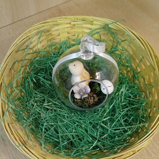 Handgefertigte, transparente Osterkugel aus Acryl mit einer Miniaturszene im Inneren: ein kleiner Osterhase sitzt auf einem Bett aus grünem Moos, daneben ein zartes Deko-Ei und feine Blüten. Die Kugel hängt an einem weißen Satinband und wirkt natürlich, frühlingshaft und elegant – ein stilvoller Osterschmuck zum Aufhängen oder Verschenken.