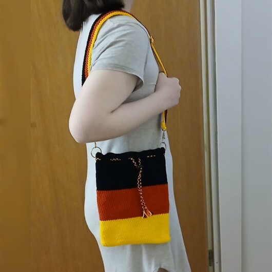 Handgehäkelte Umhängetasche in den Farben Schwarz, Rot und Gold, inspiriert von der deutschen Flagge. Mit breitem, verstellbarem Schultergurt, stabilem Kordelzugverschluss und dekorativem Kordelende. Etwa 19 x 26 cm groß, bietet Platz für Alltagsutensilien wie Handy, Geldbörse und Schlüssel. Stilvolles, handgemachtes Accessoire für Fans und Liebhaber deutscher Farben.