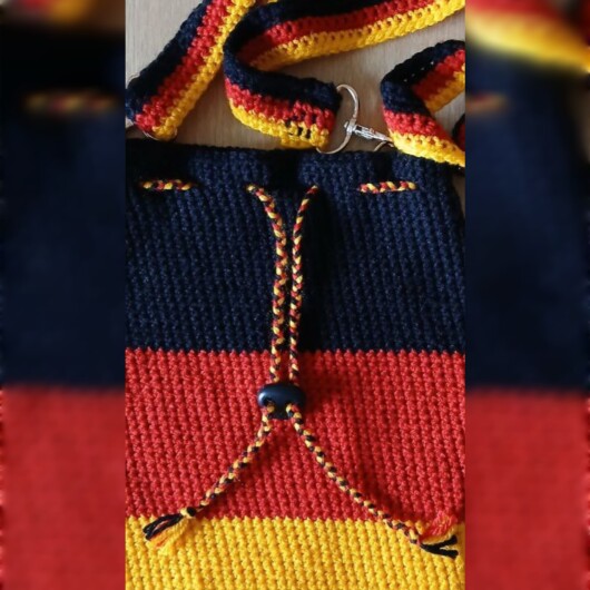 Handgehäkelte Umhängetasche in den Farben Schwarz, Rot und Gold, inspiriert von der deutschen Flagge. Mit breitem, verstellbarem Schultergurt, stabilem Kordelzugverschluss und dekorativem Kordelende. Etwa 19 x 26 cm groß, bietet Platz für Alltagsutensilien wie Handy, Geldbörse und Schlüssel. Stilvolles, handgemachtes Accessoire für Fans und Liebhaber deutscher Farben.