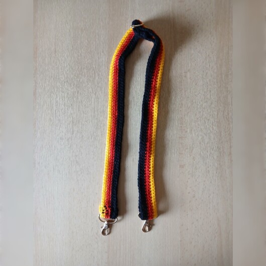Handgehäkelte Umhängetasche in den Farben Schwarz, Rot und Gold, inspiriert von der deutschen Flagge. Mit breitem, verstellbarem Schultergurt, stabilem Kordelzugverschluss und dekorativem Kordelende. Etwa 19 x 26 cm groß, bietet Platz für Alltagsutensilien wie Handy, Geldbörse und Schlüssel. Stilvolles, handgemachtes Accessoire für Fans und Liebhaber deutscher Farben.