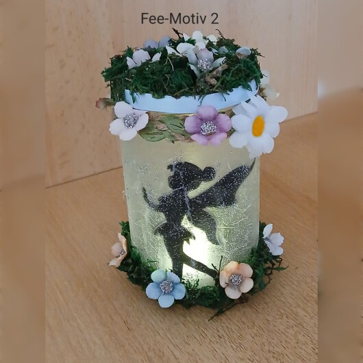 Handgefertigtes Feenglas Fee Motiv 2 mit romantischem Feenmotiv, verziert mit blauem Satinband, feinem Glitzerstaub und einer kleinen Mooslandschaft auf dem Deckel. Umgeben von zarten Kunstblumen wirkt das Glas wie ein leuchtender Miniaturgarten. Im Inneren sorgt ein flackerndes LED-Teelicht für warmes, magisches Licht – ein märchenhaftes Deko- und Nachtlicht für jeden Raum.