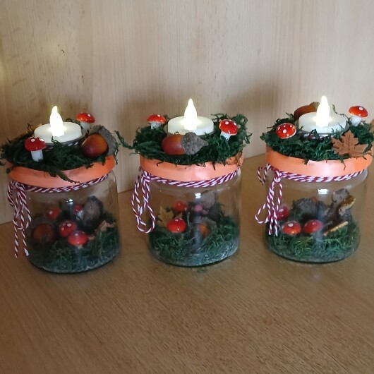 Herbstliches Windlichtglas dekoriert mit Moos, bunten Blättern, kleinen Pilzen, Eicheln und einer kleinen Igel-Figur im Inneren. Auf dem Deckel ist eine Teelichthalterung mit LED-Teelicht integriert. Der Deckel ist mit orangem Satinband verziert, das Glas zusätzlich mit rot-weißer Schnur. Stimmungsvolle Herbstdekoration für Fensterbank, Regal oder Tisch.