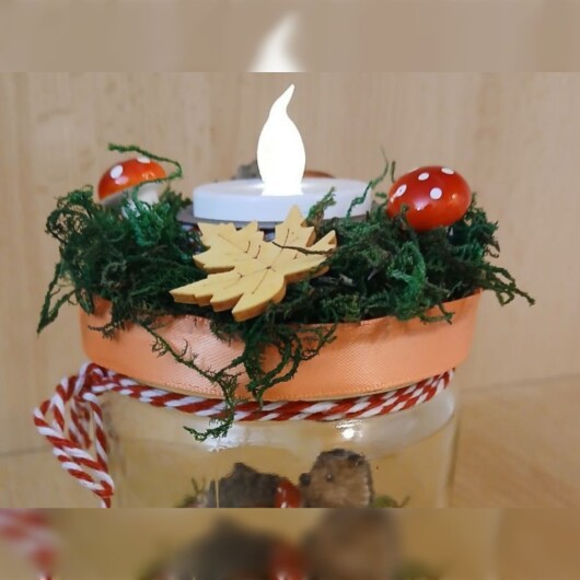 Herbstliches Windlichtglas dekoriert mit Moos, bunten Blättern, kleinen Pilzen, Eicheln und einer kleinen Igel-Figur im Inneren. Auf dem Deckel ist eine Teelichthalterung mit LED-Teelicht integriert. Der Deckel ist mit orangem Satinband verziert, das Glas zusätzlich mit rot-weißer Schnur. Stimmungsvolle Herbstdekoration für Fensterbank, Regal oder Tisch.