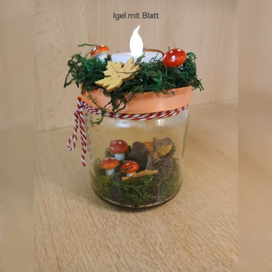 Herbstliches Windlichtglas Igel mit Blatt dekoriert mit Moos, bunten Blättern, kleinen Pilzen, Eicheln und einer kleinen Igel-Figur im Inneren. Auf dem Deckel ist eine Teelichthalterung mit LED-Teelicht integriert. Der Deckel ist mit orangem Satinband verziert, das Glas zusätzlich mit rot-weißer Schnur. Stimmungsvolle Herbstdekoration für Fensterbank, Regal oder Tisch.