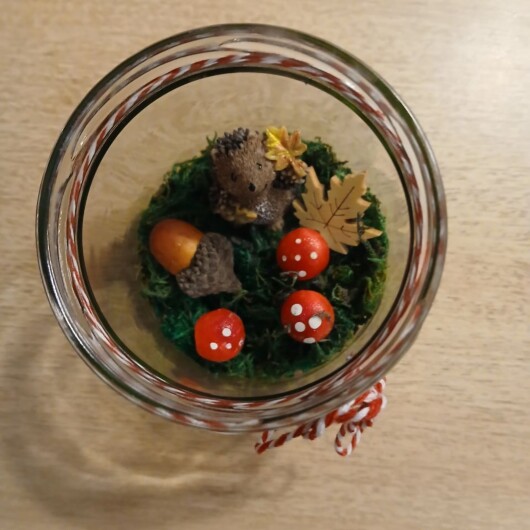 Herbstliches Windlichtglas Igel mit Blatt dekoriert mit Moos, bunten Blättern, kleinen Pilzen, Eicheln und einer kleinen Igel-Figur im Inneren. Auf dem Deckel ist eine Teelichthalterung mit LED-Teelicht integriert. Der Deckel ist mit orangem Satinband verziert, das Glas zusätzlich mit rot-weißer Schnur. Stimmungsvolle Herbstdekoration für Fensterbank, Regal oder Tisch.