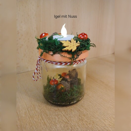 Herbstliches Windlichtglas Igel mit Nuss dekoriert mit Moos, bunten Blättern, kleinen Pilzen, Eicheln und einer kleinen Igel-Figur im Inneren. Auf dem Deckel ist eine Teelichthalterung mit LED-Teelicht integriert. Der Deckel ist mit orangem Satinband verziert, das Glas zusätzlich mit rot-weißer Schnur. Stimmungsvolle Herbstdekoration für Fensterbank, Regal oder Tisch.