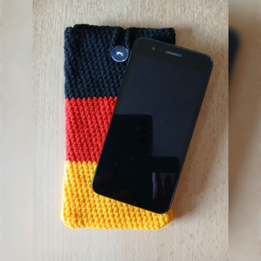 Handgehäkelte Handyhülle groß 10x18 cm in den Farben Schwarz, Rot und Gold, angeordnet wie die deutsche Flagge. Aus robustem Baumwollgarn gefertigt, mit weicher Struktur und sicherem Knopfverschluss oben. Schützt das Smartphone vor Kratzern und Stößen. Ein handgemachtes Accessoire für Fans und Liebhaber deutscher Farben.