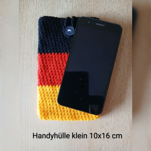 Handgehäkelte Handyhülle klein 10x16 cm in den Farben Schwarz, Rot und Gold, angeordnet wie die deutsche Flagge. Aus robustem Baumwollgarn gefertigt, mit weicher Struktur und sicherem Knopfverschluss oben. Schützt das Smartphone vor Kratzern und Stößen. Ein handgemachtes Accessoire für Fans und Liebhaber deutscher Farben.