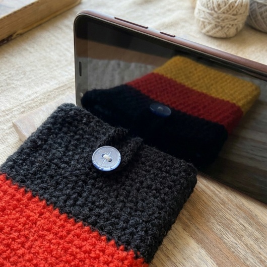 Handgehäkelte Handyhülle Deutschland Handyhülle in den Farben Schwarz, Rot und Gold, angeordnet wie die deutsche Flagge. Aus robustem Wollgarn gefertigt, mit weicher Struktur und sicherem Knopfverschluss oben. Schützt das Smartphone vor Kratzern und Stößen. Erhältlich in zwei Größen – ein handgemachtes Accessoire für Fans und Liebhaber deutscher Farben.