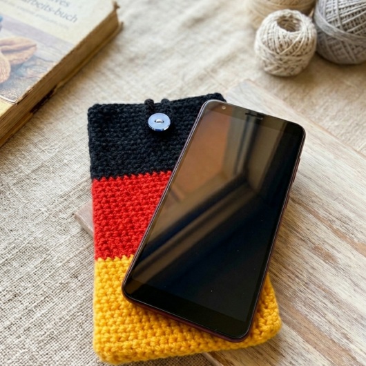 Handgehäkelte Handyhülle Deutschland Handyhülle in den Farben Schwarz, Rot und Gold, angeordnet wie die deutsche Flagge. Aus robustem Wollgarn gefertigt, mit weicher Struktur und sicherem Knopfverschluss oben. Schützt das Smartphone vor Kratzern und Stößen. Erhältlich in zwei Größen – ein handgemachtes Accessoire für Fans und Liebhaber deutscher Farben.