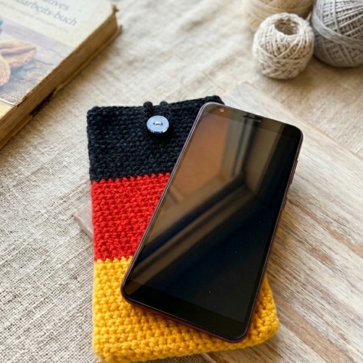 Handgehäkelte Handyhülle Deutschland Handyhülle in den Farben Schwarz, Rot und Gold, angeordnet wie die deutsche Flagge. Aus robustem Wollgarn gefertigt, mit weicher Struktur und sicherem Knopfverschluss oben. Schützt das Smartphone vor Kratzern und Stößen. Erhältlich in zwei Größen – ein handgemachtes Accessoire für Fans und Liebhaber deutscher Farben.