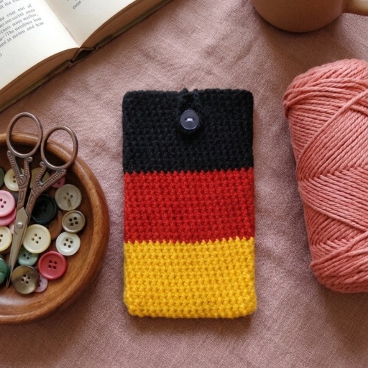 Handgehäkelte Handyhülle Deutschland Handyhülle in den Farben Schwarz, Rot und Gold, angeordnet wie die deutsche Flagge. Aus robustem Wollgarn gefertigt, mit weicher Struktur und sicherem Knopfverschluss oben. Schützt das Smartphone vor Kratzern und Stößen. Erhältlich in zwei Größen – ein handgemachtes Accessoire für Fans und Liebhaber deutscher Farben.