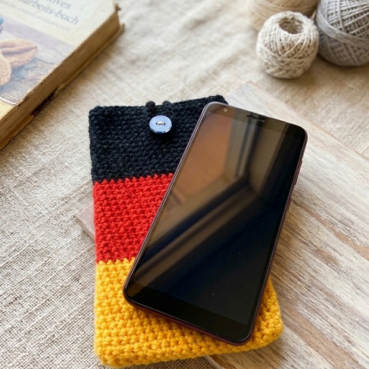 Handgehäkelte Handyhülle Deutschland Handyhülle in den Farben Schwarz, Rot und Gold, angeordnet wie die deutsche Flagge. Aus robustem Wollgarn gefertigt, mit weicher Struktur und sicherem Knopfverschluss oben. Schützt das Smartphone vor Kratzern und Stößen. Erhältlich in zwei Größen – ein handgemachtes Accessoire für Fans und Liebhaber deutscher Farben.