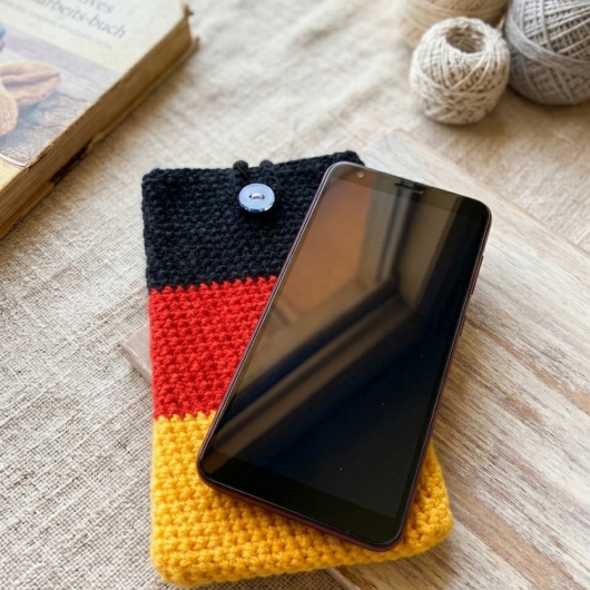 Handgehäkelte Handyhülle Deutschland Handyhülle in den Farben Schwarz, Rot und Gold, angeordnet wie die deutsche Flagge. Aus robustem Wollgarn gefertigt, mit weicher Struktur und sicherem Knopfverschluss oben. Schützt das Smartphone vor Kratzern und Stößen. Erhältlich in zwei Größen – ein handgemachtes Accessoire für Fans und Liebhaber deutscher Farben.