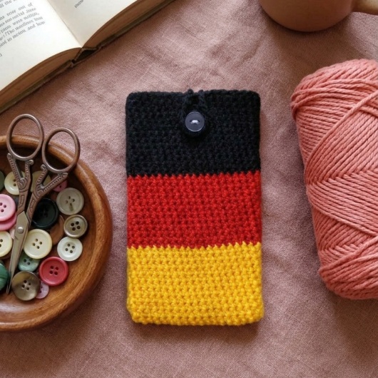 Handgehäkelte Handyhülle Deutschland Handyhülle in den Farben Schwarz, Rot und Gold, angeordnet wie die deutsche Flagge. Aus robustem Wollgarn gefertigt, mit weicher Struktur und sicherem Knopfverschluss oben. Schützt das Smartphone vor Kratzern und Stößen. Erhältlich in zwei Größen – ein handgemachtes Accessoire für Fans und Liebhaber deutscher Farben.
