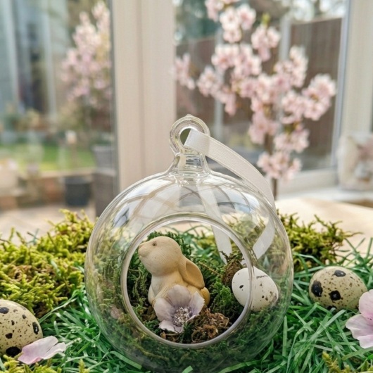 Handgefertigte Osterkugel Handgemachter Osterschmuck aus Acryl mit einer Miniaturszene im Inneren: ein kleiner Osterhase sitzt auf einem Bett aus grünem Moos, daneben ein zartes Deko-Ei und feine Blüten. Die Kugel hängt an einem weißen Satinband und wirkt natürlich, frühlingshaft und elegant – ein stilvoller Osterschmuck zum Aufhängen oder Verschenken.