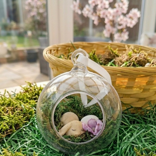 Handgefertigte Osterkugel Handgemachter Osterschmuck aus Acryl mit einer Miniaturszene im Inneren: ein kleiner Osterhase sitzt auf einem Bett aus grünem Moos, daneben ein zartes Deko-Ei und feine Blüten. Die Kugel hängt an einem weißen Satinband und wirkt natürlich, frühlingshaft und elegant – ein stilvoller Osterschmuck zum Aufhängen oder Verschenken.