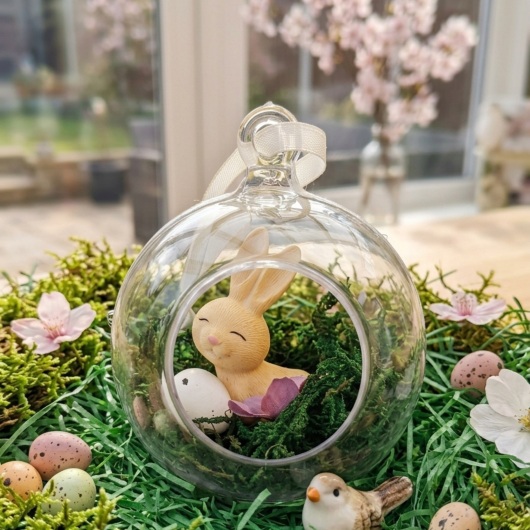 Handgefertigte Osterkugel Handgemachter Osterschmuck aus Acryl mit einer Miniaturszene im Inneren: ein kleiner Osterhase sitzt auf einem Bett aus grünem Moos, daneben ein zartes Deko-Ei und feine Blüten. Die Kugel hängt an einem weißen Satinband und wirkt natürlich, frühlingshaft und elegant – ein stilvoller Osterschmuck zum Aufhängen oder Verschenken.