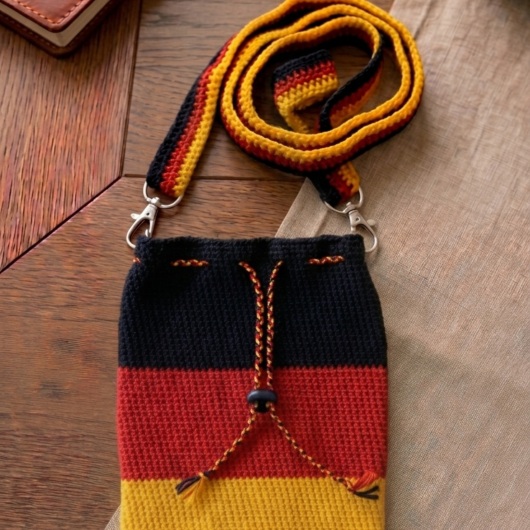 Handgehäkelte Tasche Deutschland Tasche in den Farben Schwarz, Rot und Gold, inspiriert von der deutschen Flagge. Mit breitem, verstellbarem Schultergurt, stabilem Kordelzugverschluss und dekorativem Kordelende. Etwa 19 x 26 cm groß, bietet Platz für Alltagsutensilien wie Handy, Geldbörse und Schlüssel. Stilvolles, handgemachtes Accessoire für Fans und Liebhaber deutscher Farben.