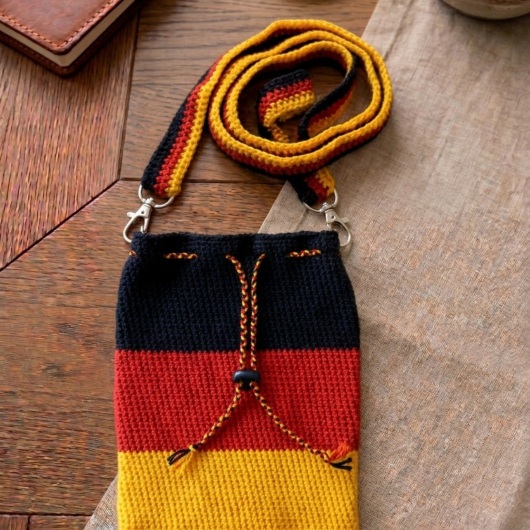 Handgehäkelte Tasche Deutschland Tasche in den Farben Schwarz, Rot und Gold, inspiriert von der deutschen Flagge. Mit breitem, verstellbarem Schultergurt, stabilem Kordelzugverschluss und dekorativem Kordelende. Etwa 19 x 26 cm groß, bietet Platz für Alltagsutensilien wie Handy, Geldbörse und Schlüssel. Stilvolles, handgemachtes Accessoire für Fans und Liebhaber deutscher Farben.