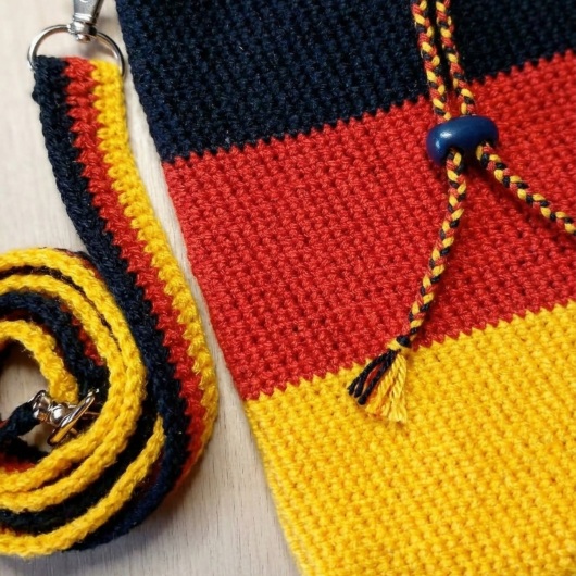 Handgehäkelte Tasche Deutschland Tasche in den Farben Schwarz, Rot und Gold, inspiriert von der deutschen Flagge. Mit breitem, verstellbarem Schultergurt, stabilem Kordelzugverschluss und dekorativem Kordelende. Etwa 19 x 26 cm groß, bietet Platz für Alltagsutensilien wie Handy, Geldbörse und Schlüssel. Stilvolles, handgemachtes Accessoire für Fans und Liebhaber deutscher Farben.