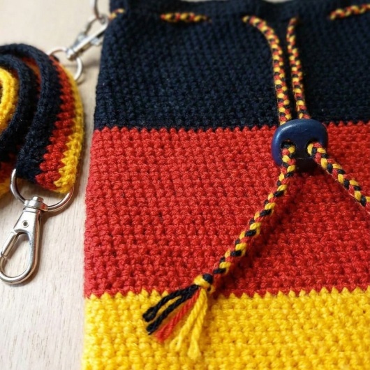 Handgehäkelte Tasche Deutschland Tasche in den Farben Schwarz, Rot und Gold, inspiriert von der deutschen Flagge. Mit breitem, verstellbarem Schultergurt, stabilem Kordelzugverschluss und dekorativem Kordelende. Etwa 19 x 26 cm groß, bietet Platz für Alltagsutensilien wie Handy, Geldbörse und Schlüssel. Stilvolles, handgemachtes Accessoire für Fans und Liebhaber deutscher Farben.