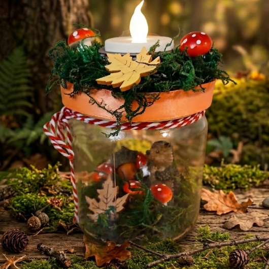 Herbstliches Kerzenglas herbstliches Windlicht dekoriert mit Moos, bunten Blättern, kleinen Pilzen, Eicheln und einer kleinen Igel-Figur im Inneren. Auf dem Deckel ist eine Teelichthalterung mit LED-Teelicht integriert. Der Deckel ist mit orangem Satinband verziert, das Glas zusätzlich mit rot-weißer Schnur. Stimmungsvolle Herbstdekoration für Fensterbank, Regal oder Tisch.